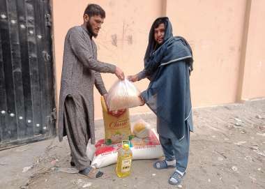 Left: a young man in beige Islamic dress hands a bag of food supplies to a man on the right, wearing blue clothes and head covering.